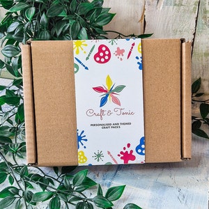 Puede incluir: Una caja de cart&oacute;n marr&oacute;n con una etiqueta colorida que dice "Craft & Tonic" y "Packs de manualidades personalizados y tem&aacute;ticos". La etiqueta tiene un dise&ntilde;o de pinceles, salpicaduras de pintura y una paleta de pintura.