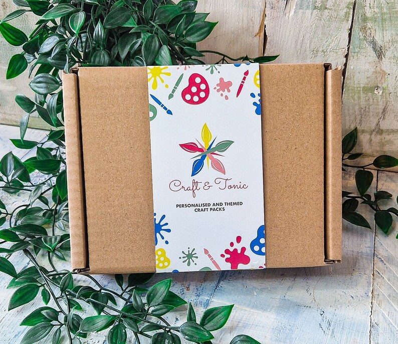 Puede incluir: Una caja de cart&oacute;n marr&oacute;n con una etiqueta colorida que dice "Craft & Tonic" y "Packs de manualidades personalizados y tem&aacute;ticos". La etiqueta tiene un dise&ntilde;o de pinceles, salpicaduras de pintura y una paleta de pintura.