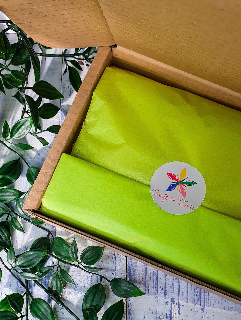 May include: A cardboard box with a green tissue paper lining. The box is open and the tissue paper is visible. A white circle sticker with the text "Craft & Tonic" and a colorful flower design is on the tissue paper.