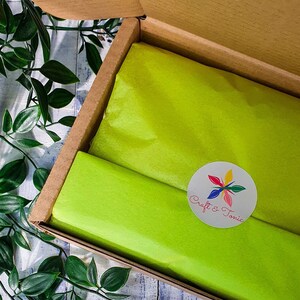Puede incluir: Una caja de cart&oacute;n con un forro de papel de seda verde. El papel de seda tiene una pegatina circular blanca con un dise&ntilde;o de flor colorido y el texto "Craft & Tonic".