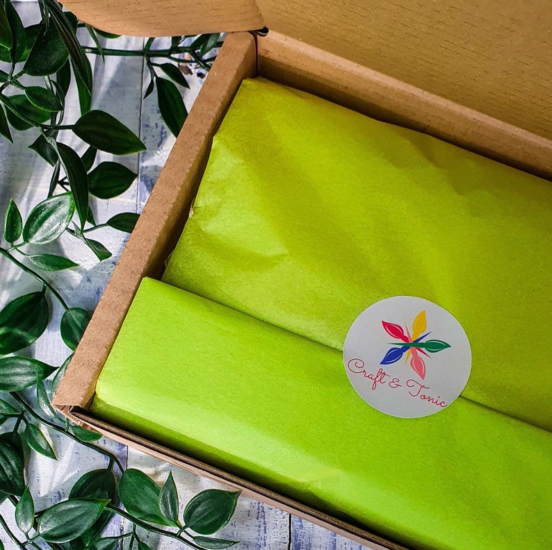 Puede incluir: Una caja de cart&oacute;n con un forro de papel de seda verde. El papel de seda tiene una pegatina circular blanca con un dise&ntilde;o de flor colorido y el texto "Craft & Tonic".