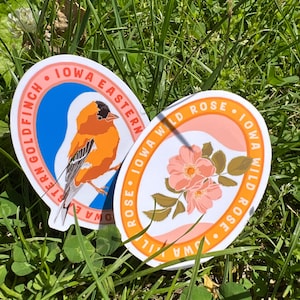 May include: Two oval stickers featuring Iowa state symbols. One shows an orange and black Eastern Goldfinch with the text "IOWA EASTERN GOLDFINCH". The other depicts a pink Iowa Wild Rose with the text "IOWA WILD ROSE".