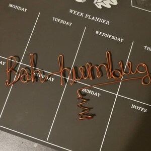 May include: A copper-colored wire sculpture formed into the cursive text "Bah humbug" with a coiled base. The wire is shaped into a continuous line, creating the letters of the phrase, and features a spiral stand at the bottom for stability.