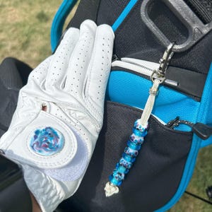 May include: A white golf glove with a circular blue, purple, and white marbled accent on the wrist strap, paired with a matching beaded zipper pull featuring blue and white patterned glass beads on a white cord, attached to a black and blue golf bag.