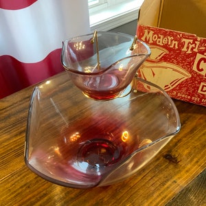 May include: A vintage three-piece glass serving set with a clear glass bowl and a smaller bowl with a pink tint. The set is made of glass and has a gold metal stand. The box reads "Modern Tri-Level Chip & Dip Set".