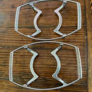 May include: Two silver metal hot pad trivets with a wavy design. The trivets are oval shaped and have a raised edge.