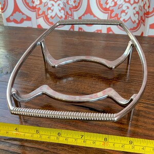 May include: A silver metal trivet with a wavy design and a beaded edge. The trivet measures approximately 8 inches in length.