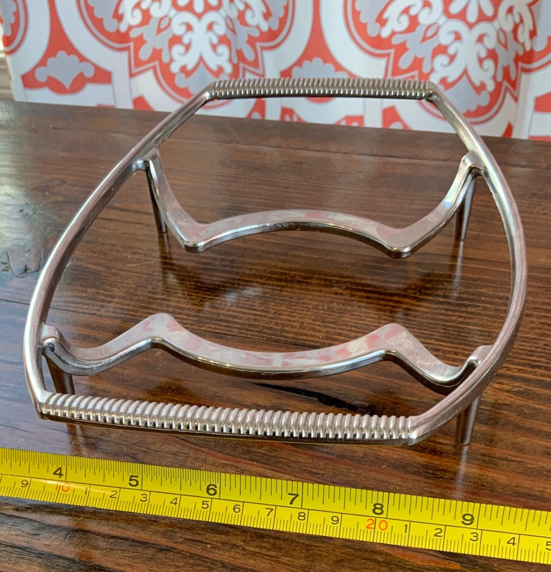 May include: A silver metal trivet with a wavy design and a beaded edge. The trivet measures approximately 8 inches in length.