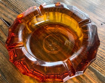 May include: Amber glass ashtray with a faceted design. The ashtray has a circular shape with a deep well in the center.