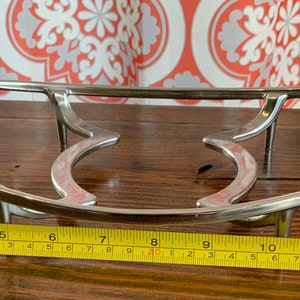 May include: A silver metal hot plate stand with a curved design and three legs. The stand measures 28 cm in length.