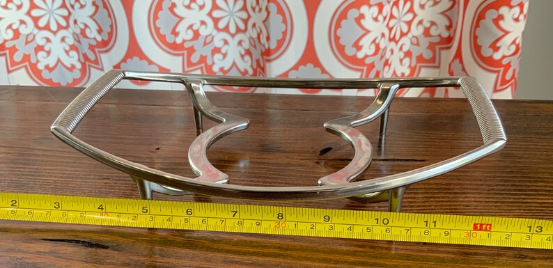 May include: A silver metal hot plate stand with a curved design and three legs. The stand measures 28 cm in length.