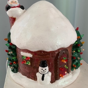 May include: A ceramic Christmas village scene with a brown brick house, a white snow-covered roof, a snowman with a black top hat and tie, and a Santa Claus figure on the chimney. The house has a red door and windows with colorful lights.
