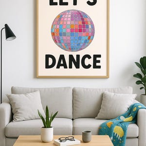 Op de afbeelding: Een ingelijste print met de woorden "LET'S DANCE" in zwarte blokletters. Het kunstwerk toont een kleurrijke discobal-afbeelding. De print wordt weergegeven boven een lichtgrijze bank met kussens en een houten salontafel.