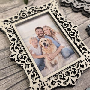 May include: Ornate, cream-colored picture frame with a family photo. The frame has a decorative, openwork design. The photo shows a smiling family with a golden retriever dog. The frame is on a wooden surface.
