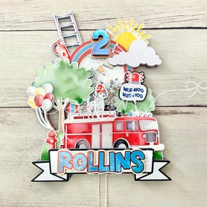 May include: A colorful, layered cake topper featuring a red fire truck, a Dalmatian, and the number 2. Includes a rainbow, sun, clouds, and the name "ROLLINS" on a banner. Other elements include a ladder, fire hydrant, and balloons.