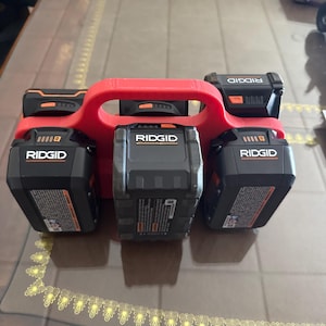 Op de afbeelding: Een rode, 3D-geprinte batterijhouder met vijf zwarte en oranje Ridgid-accu's voor elektrisch gereedschap. De batterijen zijn gerangschikt in een houder met een handvat voor gemakkelijk dragen. De batterijen hebben het Ridgid-logo en productinformatie.