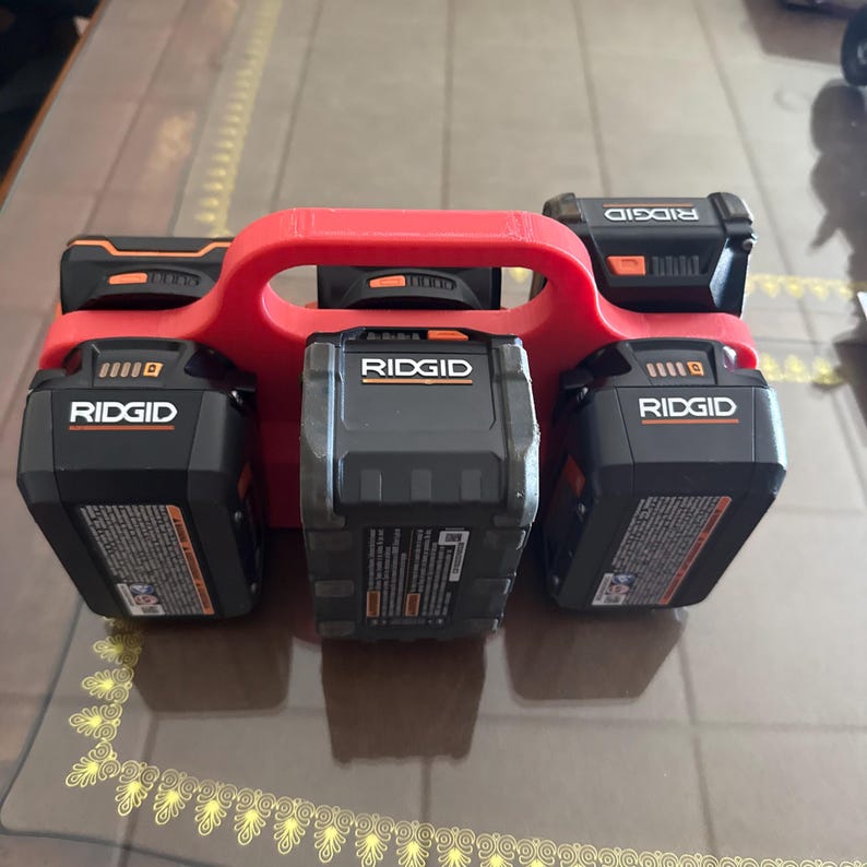 Op de afbeelding: Een rode, 3D-geprinte batterijhouder met vijf zwarte en oranje Ridgid-accu's voor elektrisch gereedschap. De batterijen zijn gerangschikt in een houder met een handvat voor gemakkelijk dragen. De batterijen hebben het Ridgid-logo en productinformatie.