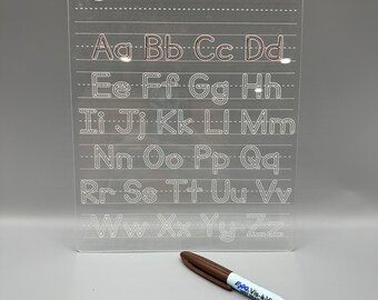May include: A clear plastic dry-erase board with a grid of lines and dotted letters of the alphabet, 'Aa Bb Cc Dd Ee Ff Gg Hh Ii Jj Kk Ll Mm Nn Oo Pp Qq Rr Ss Tt Uu Vv Ww Xx Yy Zz', for practicing handwriting. A brown dry-erase marker is lying next to the board.