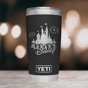 May include: Black YETI travel mug featuring a silver Disney castle and fireworks design. The Disney logo is below the castle. The mug has a silver rim and base, and the YETI logo is at the bottom.
