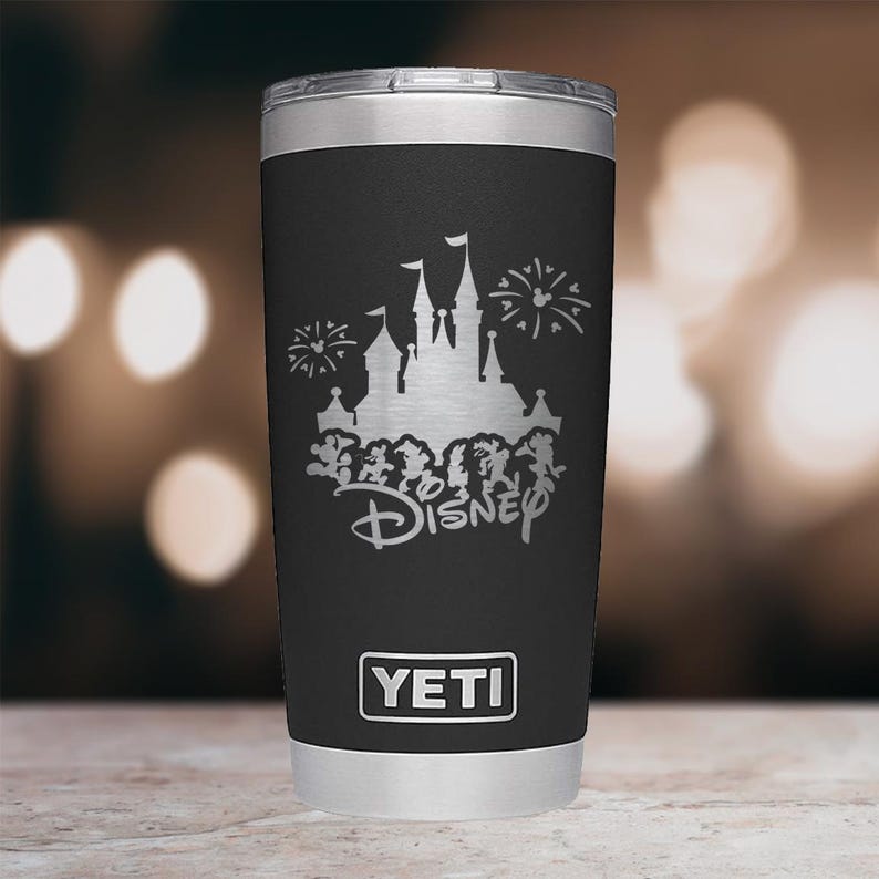 May include: Black YETI travel mug featuring a silver Disney castle and fireworks design. The Disney logo is below the castle. The mug has a silver rim and base, and the YETI logo is at the bottom.