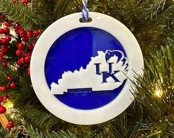 May include: A white and blue Christmas ornament featuring the outline of the state of Kentucky with the letters "UK" and the word "Wildcats" inside. The ornament is hanging on a Christmas tree.