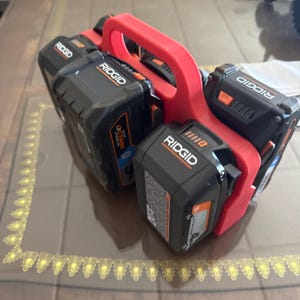 Op de afbeelding: Een rode 3D-geprinte batterijhouder met vier zwarte en oranje Ridgid-elektrische gereedschapsbatterijen. De batterijen zijn in een compacte configuratie gerangschikt, met het Ridgid-logo prominent aanwezig.