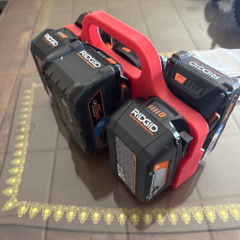 Op de afbeelding: Een rode 3D-geprinte batterijhouder met vier zwarte en oranje Ridgid-elektrische gereedschapsbatterijen. De batterijen zijn in een compacte configuratie gerangschikt, met het Ridgid-logo prominent aanwezig.