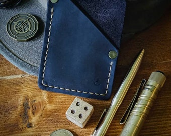 Peut inclure: Un portefeuille en cuir noir avec un bord cousu, une boussole, un dé, une pièce de monnaie, un stylo et une lampe de poche sur une surface en bois.