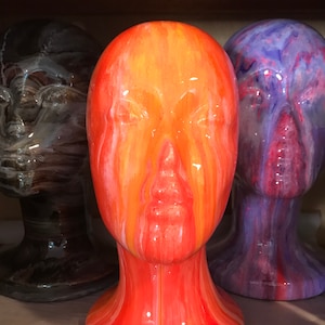 May include: Three ceramic mannequin heads with abstract paint designs. The head in the centre is orange and yellow with white streaks. The head on the left is brown and white. The head on the right is purple and pink with white streaks.
