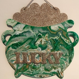 Peut inclure: Un pot d'or vert et or avec le mot "lucky" écrit en paillettes dorées. Le pot est décoré d'un motif de peinture tourbillonnant vert et blanc.
