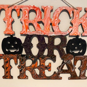 May include: A wooden sign with the words "Trick or Treat" painted in orange, black and white. The letters are outlined in black and have a marbled effect. Two black silhouettes of jack-o'-lanterns are placed between the words "or" and "treat".