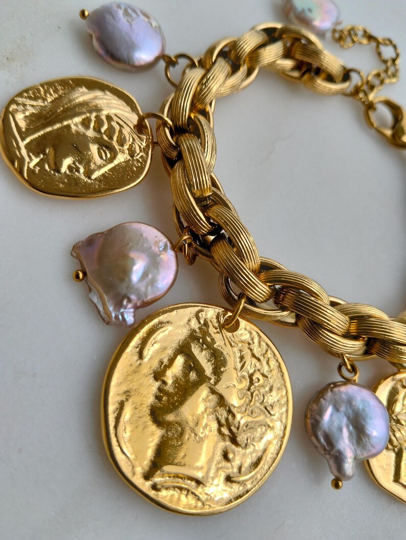 May include: Gold-toned bracelet featuring a thick chain design, adorned with coin-shaped charms and irregularly shaped, iridescent pearls. The charms depict classical figures. The bracelet is displayed on a white surface.