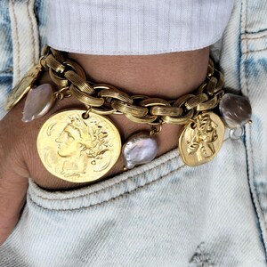 May include: Gold-toned charm bracelet featuring a chunky chain design. The bracelet is adorned with coin-shaped charms and irregularly shaped pearls. The bracelet is worn on a wrist, with a white shirt and denim jacket in the background.