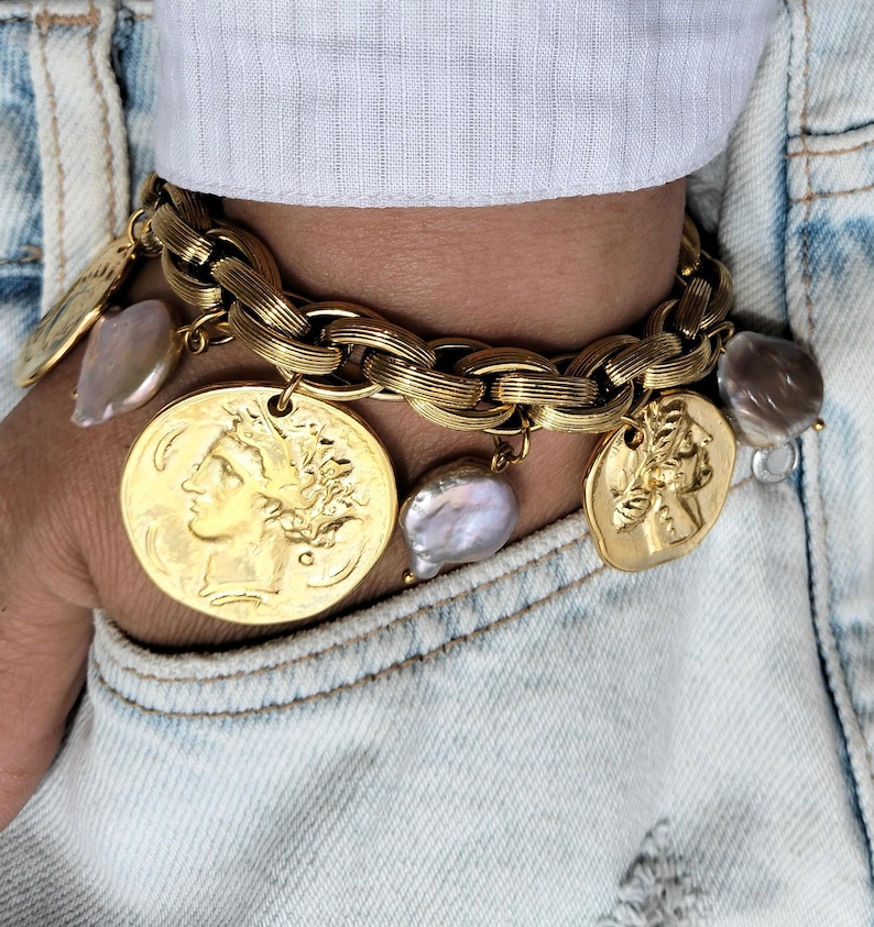 May include: Gold-toned charm bracelet featuring a chunky chain design. The bracelet is adorned with coin-shaped charms and irregularly shaped pearls. The bracelet is worn on a wrist, with a white shirt and denim jacket in the background.