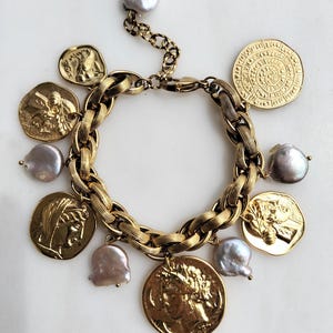 May include: A gold-toned bracelet featuring a chunky chain design, adorned with coin charms and irregular-shaped pearls. The coins display detailed relief patterns. The bracelet is displayed on a white surface.