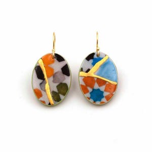 Op de afbeelding: Ovale oorbellen met kleurrijke mozaïekontwerpen. Elke oorbel heeft een uniek patroon van oranje, zwarte, blauwe en groene vormen, geaccentueerd met gouden lijnen. De oorbellen hebben goudkleurige haken en een lichtgroene rand. Ongeveer 3 cm lang.
