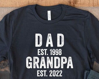 May include: Black t-shirt with white text that reads "DAD EST. 1998 GRANDPA EST. 2022". The shirt is laid out on a wooden surface with a pair of white sneakers and blue jeans.