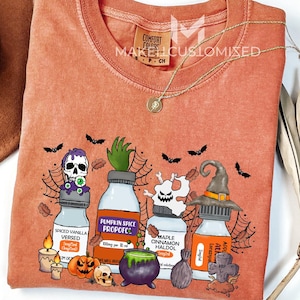 Peut inclure: T-shirt orange à col rond avec un motif sur le thème d'Halloween. Le motif présente des flacons de potion avec des étiquettes telles que "Spiced Vanilla Versed" et "Pumpkin Spice Propofc", ainsi que des chauves-souris, des crânes et un chapeau de sorcière. Un chapeau marron et un collier doré sont également visibles.