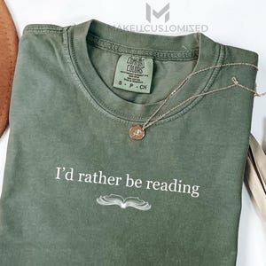 May include: An olive green t-shirt with the text "I'd rather be reading" and an open book illustration. A gold necklace with a pendant is on the shirt. A brown hat and dried plant are in the background.