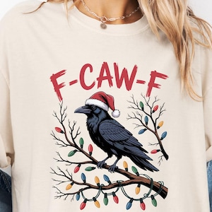 Puede incluir: Camiseta color crema con un cuervo negro con gorro de Papá Noel, posado en una rama decorada con luces navideñas. El texto "F-CAW-F" está encima del cuervo. La camiseta es de corte holgado.
