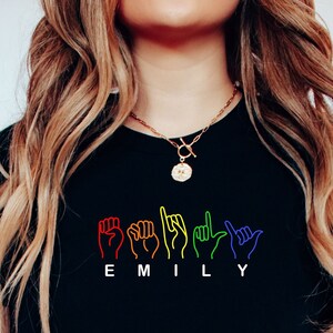 May include: A black t-shirt with a rainbow design of hands spelling the name "EMILY" in British Sign Language.