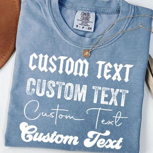 May include: A blue Comfort Colors t-shirt with the words "CUSTOM TEXT" printed in white in various fonts. A gold necklace with a pendant rests on the shirt. A brown hat and dried plant are in the background.