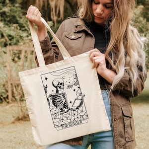 May include: A white canvas tote bag featuring a black and white illustration of a skeleton reading a book. The illustration is surrounded by flowers and stars. The text "THE READER" is printed below the illustration.