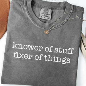 Peut inclure: Un t-shirt gris Comfort Colors avec le texte "knower of stuff fixer of things" en blanc. Un collier en or avec un pendentif est drapé sur le t-shirt. Un chapeau marron et une plante séchée sont en arrière-plan.