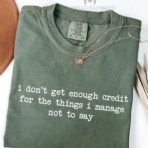 Puede incluir: Una camiseta verde oliva con el texto "i don't get enough credit for the things i manage not to say" en blanco. Un collar dorado con un colgante está drapeado sobre la camiseta. La camiseta está doblada sobre una superficie blanca.