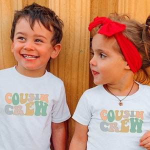 May include: Two children wearing white t-shirts with the text "Cousin Crew" in colourful retro font. The girl is wearing a red headband, a plaid shirt tied around her waist, and denim shorts. The boy is wearing blue jeans. They are standing in front of a wooden fence.