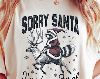May include: A cream coloured t-shirt with a black graphic of a raccoon dressed as Santa Claus riding a reindeer. The text "Sorry Santa I've been feral" is printed below the graphic.