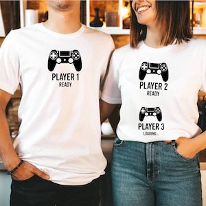 May include: Two white t-shirts with black text and a black video game controller graphic. The text on the first shirt reads "Player 1 Ready". The text on the second shirt reads "Player 2 Ready" and "Player 3 Loading..."