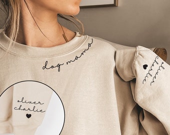May include: A beige sweatshirt with the text "dog mama" on the front, "Oliver Charlie" with a heart on the left sleeve, and "Oliver Charlie" on the right sleeve.