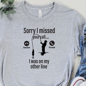 May include: Gray t-shirt with a fishing-themed design. The text reads "Sorry I missed your call... Decline Accept I was on my other line". A silhouette of a person fishing is shown with a phone icon on a fishing line.